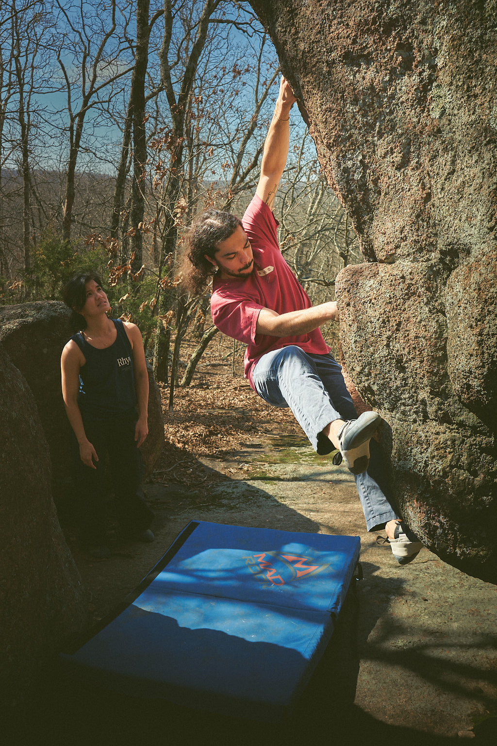 Person rock climbing in a Rhyo / 02: Process t-shirt with another person observing in a forest setting.