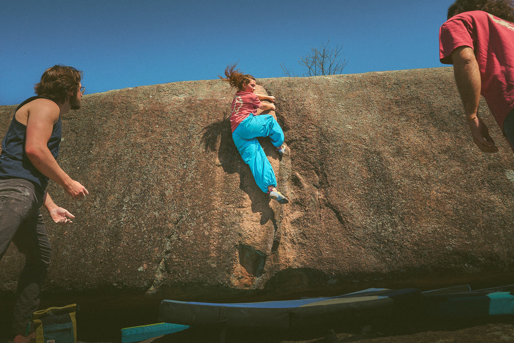 Person wearing the Rhyo / 02: Process t-shirt while climbing a large rock formation with two others assisting.